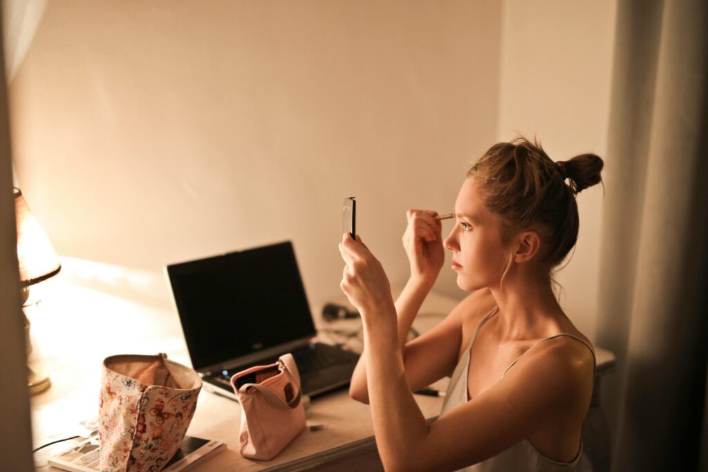 Woman applying makeup at a desk in warm lighting, with a laptop and cosmetic bags visible in the background.