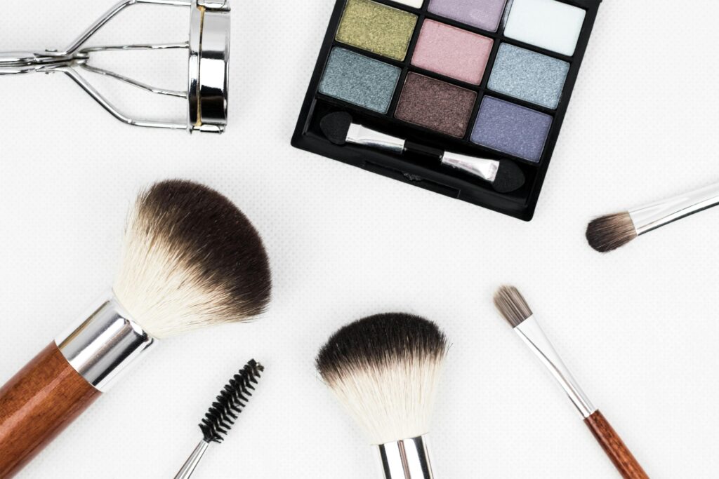 Flat lay of makeup tools including an eyeshadow palette, wooden-handled brushes, mascara wand, and metal eyelash curler.