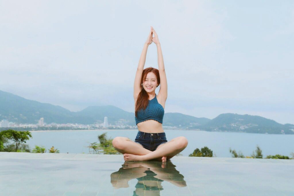 Smiling woman in a blue top practicing a seated yoga pose with raised arms at the edge of an ocean view pool.