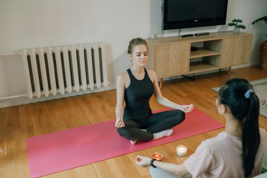 Two women meditating in Lotus position on a pink yoga mat, with lit candles between them in a living room.
