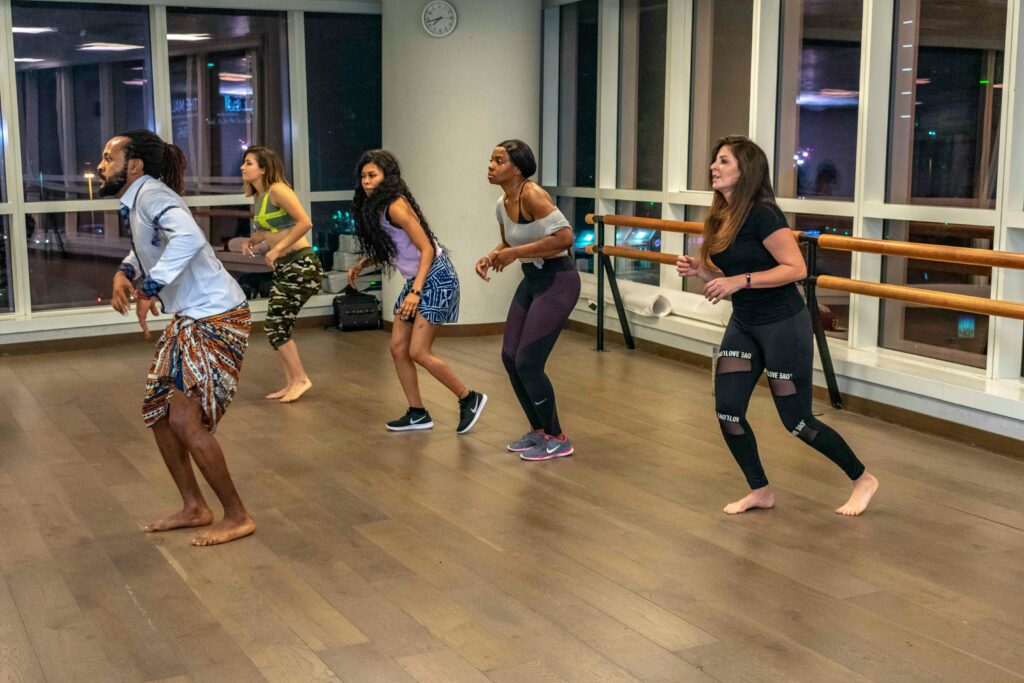 Diverse group practicing high-energy ethnic or cultural dance moves in a sleek studio overlooking a city at night.