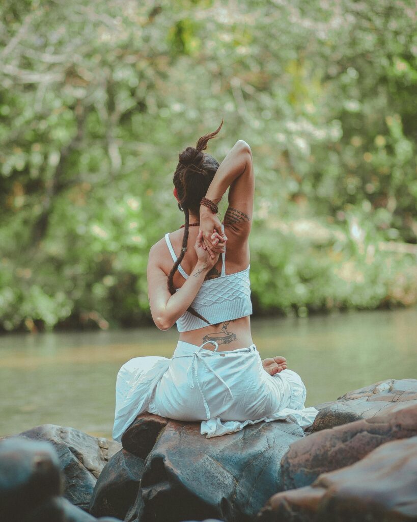 Person with dreadlocks doing a deep shoulder stretch (Gomukhasana arms) while meditating on a rock by a river.
