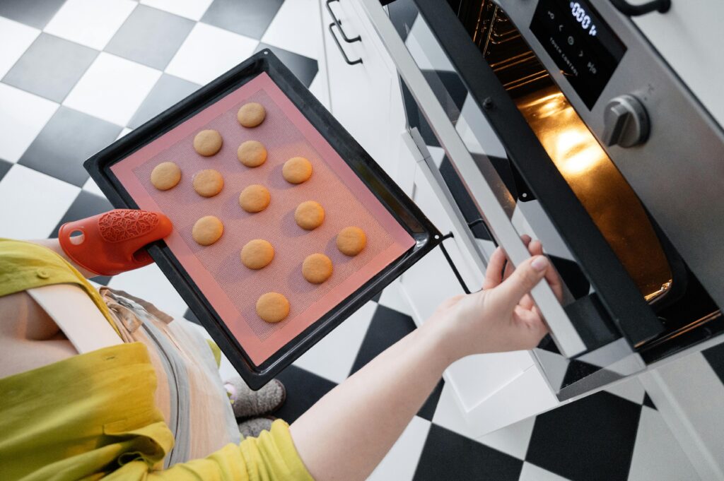 Person placing a tray of unbaked cookies into an oven in a kitchen with black and white checkered floor tiles.