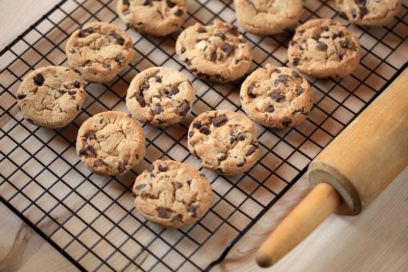 Freshly baked chocolate chip cookies cooling on a wire rack next to a wooden rolling pin on a wooden surface.