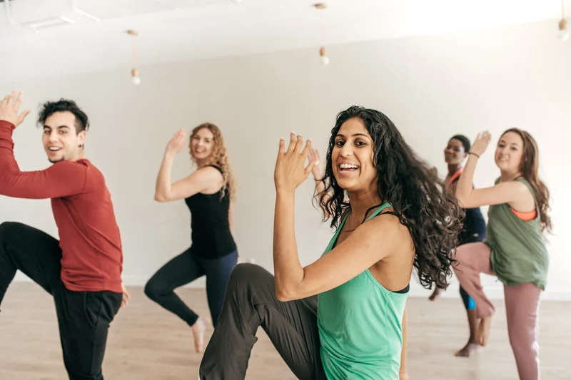 A diverse group of smiling adults performing a high-intensity step or dance aerobics workout in a bright studio.