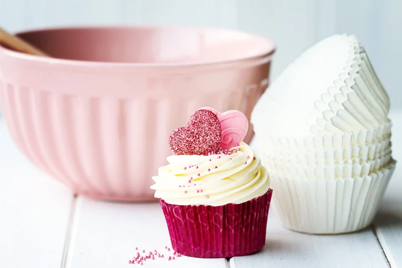 Decorated cupcake in a pink wrapper with heart-shaped sprinkles and icing, placed in front of a pink mixing bowl and a stack of white cupcake liners on a white table